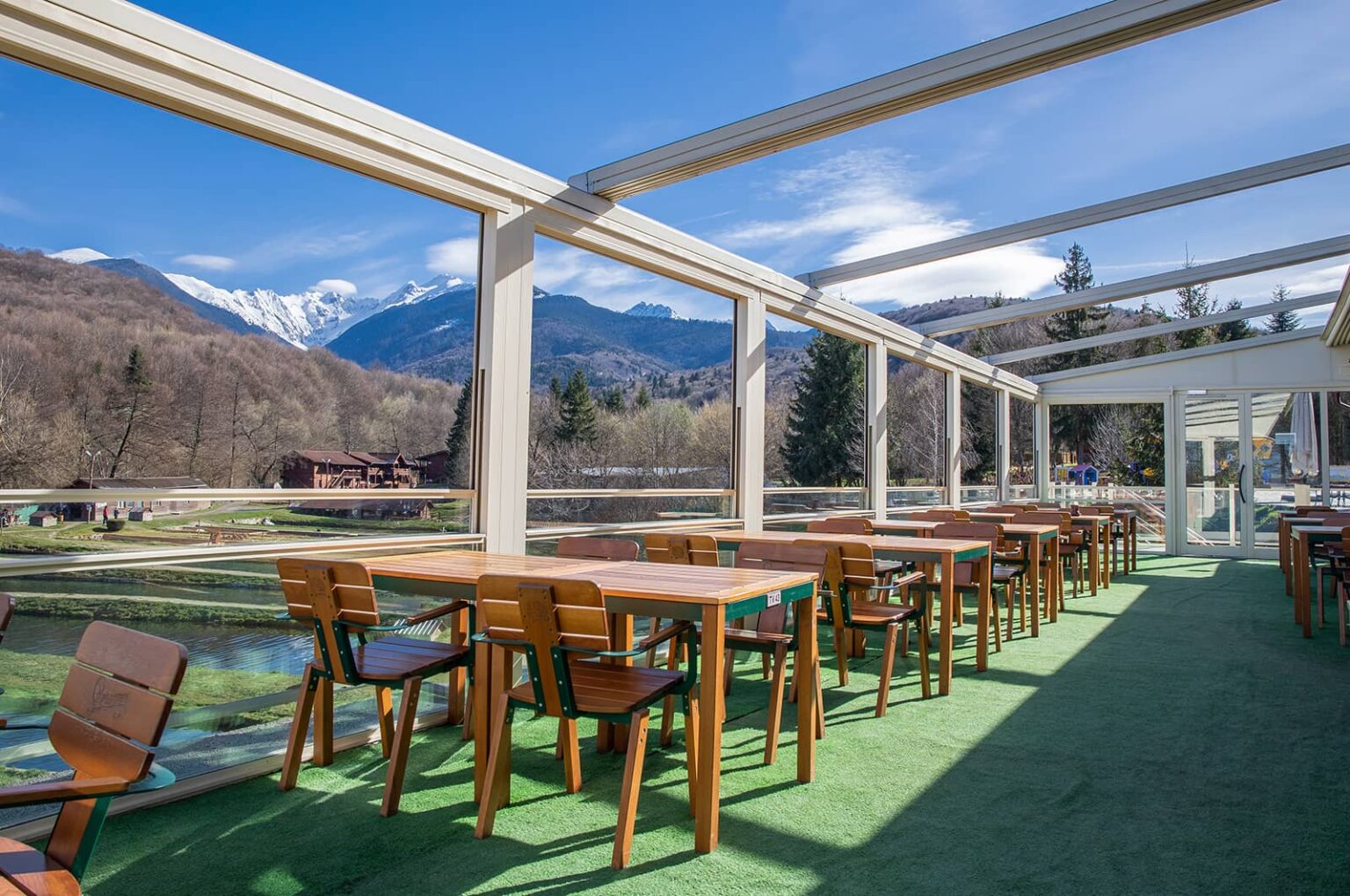 Veranda de sticlă cu mese lungi din lemn și vedere spre munții înzăpeziți