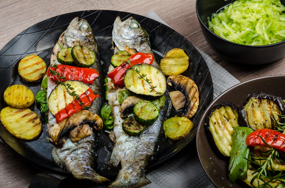 Two grilled trout with vegetables on a black tray